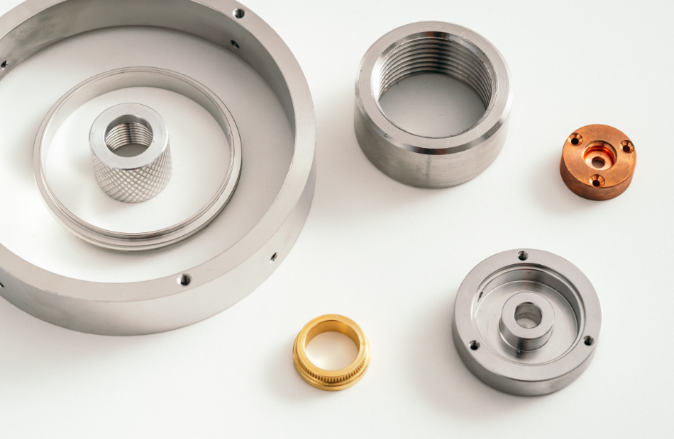 Mehrere runde Metallkomponenten unterschiedlicher Größe liegen auf einer weißen Oberfläche. Die Bauteile, darunter eine gerändelte Hülse, ein großer Ring und kleinere Drehteile aus Edelstahl und Messing, wurden durch CNC-Drehen hergestellt und weisen präzise Gewinde und Oberflächen auf.
