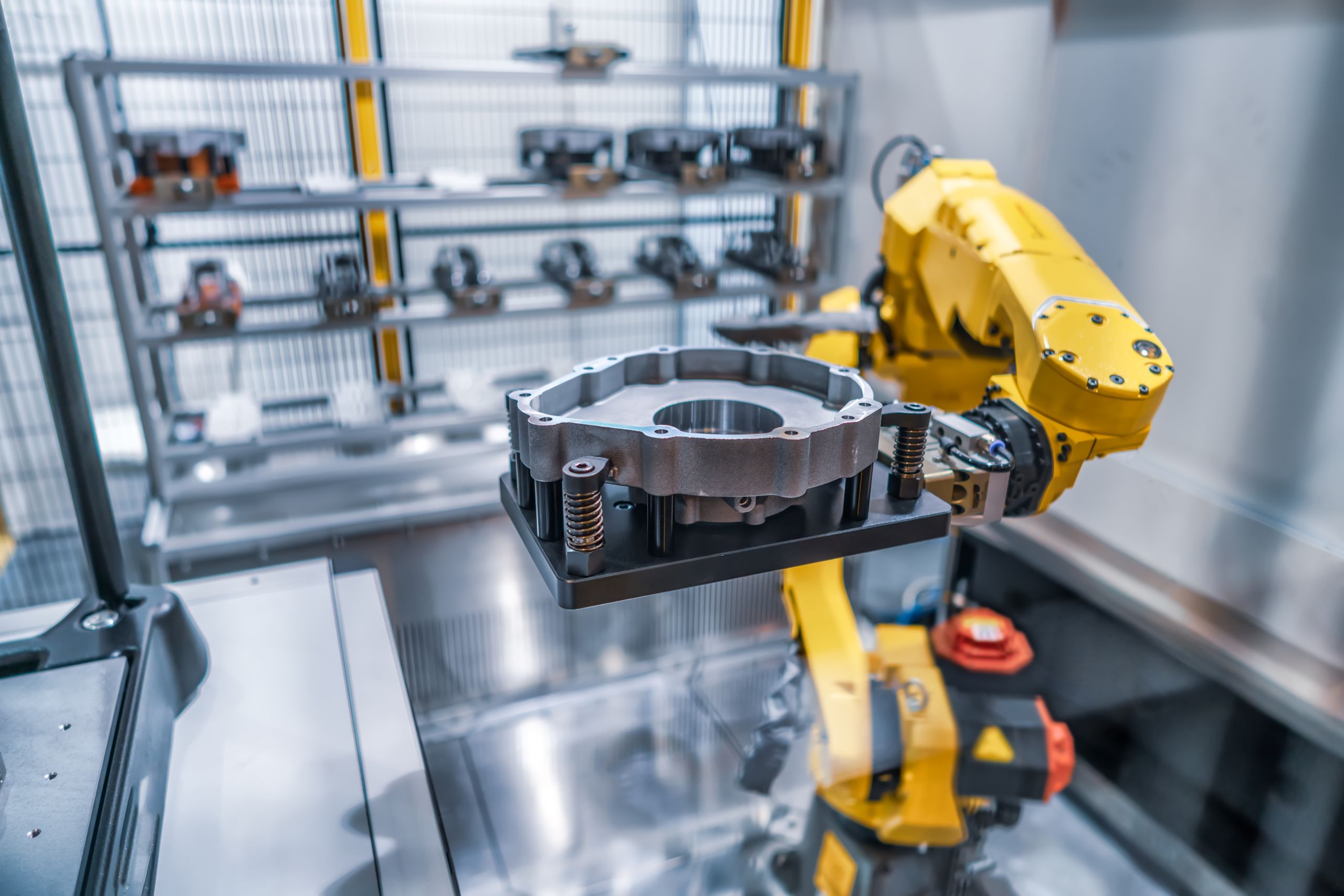 Ein gelber Roboterarm hält ein präzisionsgefertigtes Metallbauteil in einer industriellen Produktionsumgebung, mit Regalen voller Bauteile im Hintergrund.