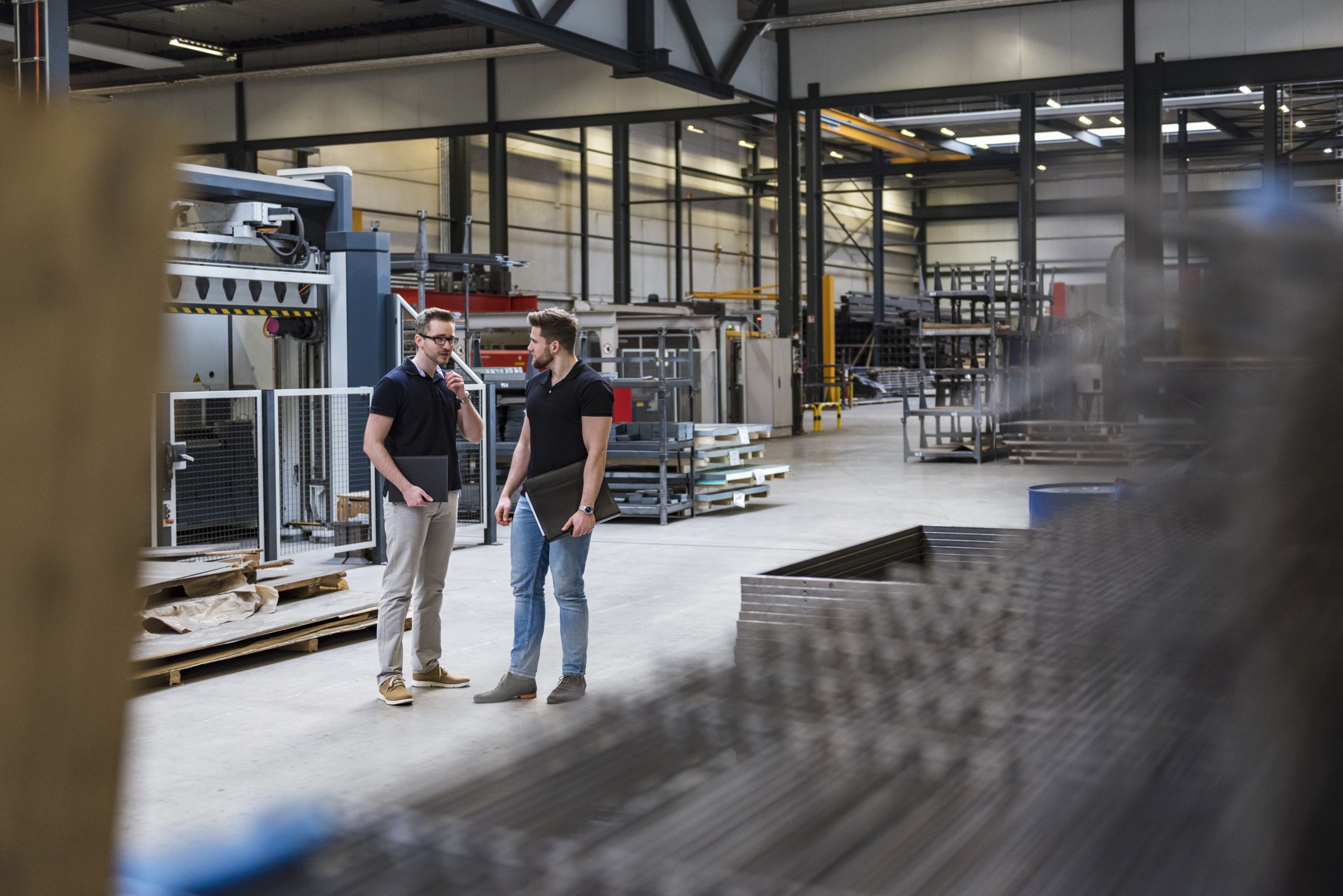 Zwei Mitarbeiter in einer Fabrikhalle, die eine Diskussion führen, während sie Laptops in der Hand halten.